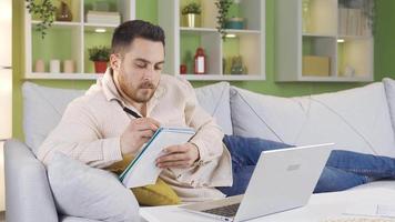 Man lying on sofa working from laptop and taking notes of what he sees.    Man thoughtfully doing his job and taking notes using laptop in living room. video