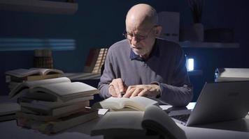 Alten Forscher oder Historiker Mann lesen ein Buch, mit Laptop allein im dunkel Studie Zimmer. nachdenklich Alten Forscher mit Laptop und Surfen wissenschaftlich Bücher im seine Büro. video