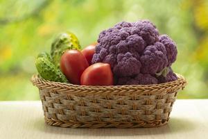 Fresh ripe purple cauliflower, red tomatoes and green cucumber in basket. Healthy food on table on defocus autumn background. photo