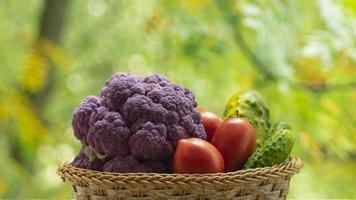 Fresh ripe purple cauliflower, red tomatoes and green cucumber in basket. Healthy food on table on defocus autumn background. photo