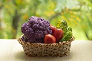 Fresh ripe purple cauliflower, red tomatoes and green cucumber in basket. Healthy food on table on defocus autumn background. photo