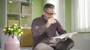le vieux homme est en train de lire une livre dans le sien confortable et Frais Accueil pendant le jour. âge moyen homme dans des lunettes en train de lire une livre dans le sien paisible moderne maison, profiter le sien gratuit temps. video
