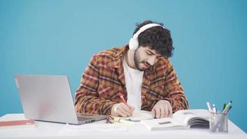 Universidad estudiante haciendo deberes a el escritorio, escuchando a música y estudiando con placer. contento joven hombre estudiando, utilizando su computadora portátil, tomando notas y escuchando a música con auriculares. video