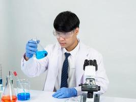 estudiante científico asiático en un laboratorio de mezcla de reactivos en un laboratorio de investigación científica con tubos de ensayo de varios tamaños y microscopios. sobre la mesa en el fondo blanco de laboratorio de química de laboratorio. foto