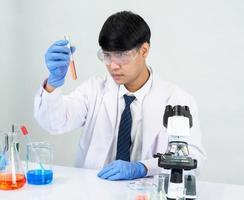 estudiante científico asiático en un laboratorio de mezcla de reactivos en un laboratorio de investigación científica con tubos de ensayo de varios tamaños y microscopios. sobre la mesa en el fondo blanco de laboratorio de química de laboratorio. foto