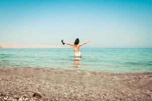 Young carefree Caucasian woman tourist dance on the beach in turquoise water alone with hands up hold slippers on summer vacation beach holidays. Solo travel adventure wanderlust photo