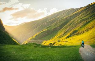 Cyclist back view on the road in green caucasus nature with scenic sunset background on valley. Traveller on bicycle. Solo travel long distance bicycle touring. Off-road biking photo