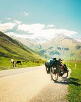 Red touring bicycle loaded with heavy gear in four pannier bags stand on side of asphalt road surrounded by summer nature and caucasus mountains background. Solo adventure travel photo
