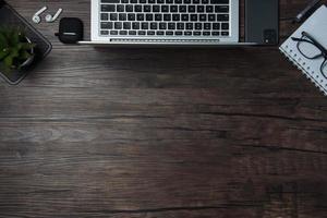 wooden office desk with laptop, blank notebook, cordless earphones and glasses, dark background, wooden background, Top view with copy space, empty space. photo