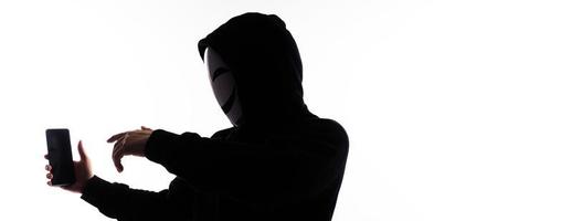 Hacker Anonymous and face mask with smartphone in hand. Man in black hood shirt holding and using mobile phone on white background. Represent cyber crime data hacking or stealing personal data concept photo