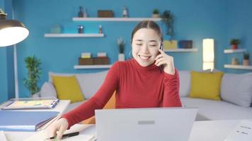 Asian woman working from home also happily chatting on the phone in a good mood. Asian young woman working from laptop at home also having nice phone call, happy. video