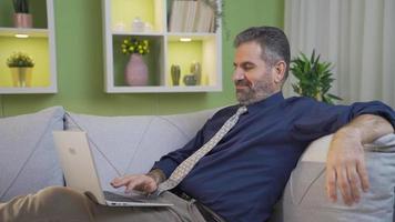 man använder sig av bärbar dator på soffa avkopplande på Hem. mogna man avkopplande på Hem liggande på soffa, använder sig av bärbar dator, skriver använder sig av tangentbord. video