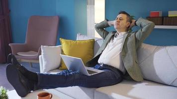 gelukkig Mens resting en vinden vrede Bij zorgeloos kalmte huis. ontspannen en zorgeloos volwassen zakenman ontspannende Aan sofa Bij huis, werken van laptop. video