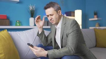 Happy adult man sitting on sofa at home using useful mobile apps. Happy man spends time alone at home with his phone, uses mobile apps, relaxes, rests. video