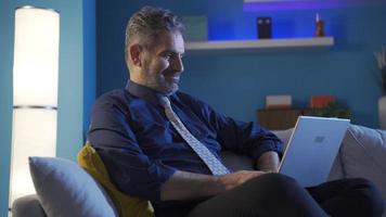 Mature businessman relaxing on sofa working on laptop, taking notes in his diary. Relaxed and peaceful businessman resting at night alone on sofa at home, doing his work with calm and relaxed head. video