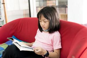 Little asian girl is reading a book while sitting near window photo
