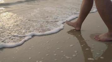 Bare feet of a man walking on the sand of a seashore. A sea wave with foam washes the bare feet of a man walking slowly along the sand. video