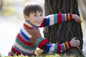 A little boy in a knitted sweater sits near a tree and looks at the camera. photo