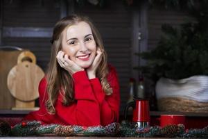 retrato de un joven rubia mujer en un rojo suéter en contra el fondo de un cocina con Navidad decoraciones veinticinco años mujer en el Mañana con un café maceta y un taza. foto