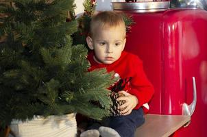 Sad little boy at christmas time. Baby is sitting on the table in the kitchen near the Christmas tree. photo