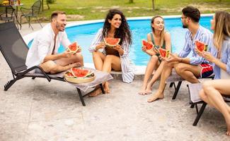 jóvenes sentados junto a la piscina y comiendo sandía en el patio trasero de la casa foto