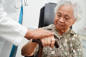 Doctor help Asian elderly disability woman patient holding walking stick in wrinkled hand at hospital. photo