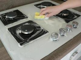 Cleaning dirty gas stove, foam from household chemicals. Hands hold a washing sponge in the foam. White glass hob surface. The concept of household chores, house cleaning. photo