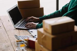 Emprendedora de pequeñas empresas o mujer asiática independiente que usa una computadora portátil con caja, joven mujer asiática de éxito con su mano levantada, caja de embalaje de marketing en línea y entrega, concepto de pyme. foto