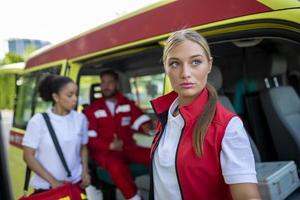 Tres multirracial paramédicos en pie en frente de ambulancia vehículo, que lleva portátil equipo foto