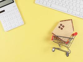 plano diseño de de madera casa modelo en compras carretilla, blanco calculadora y computadora teclado en amarillo antecedentes con Copiar espacio, hogar compra concepto. foto