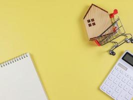 flat layout of wooden house model in shopping trolley, white calculator, blank page opened  notebook  on yellow  background with copy space, home purchase concept. photo