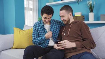 Fun and cheerful brothers looking at phone together at home. Younger brother at home with his older brother looking at the phone, surfing social media, checking posts. video