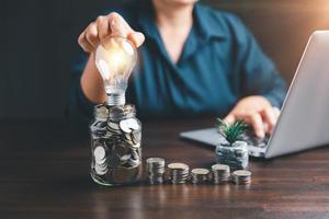 Businesswoman with lightbulb on money jar and using calculator to calculate and money stack. Save energy and money with accounting finance in office concept. Idea of energy saving planning in home. photo