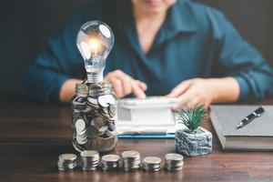 Businesswoman with lightbulb on money jar and using calculator to calculate and money stack. Save energy and money with accounting finance in office concept. Idea of energy saving planning in home. photo