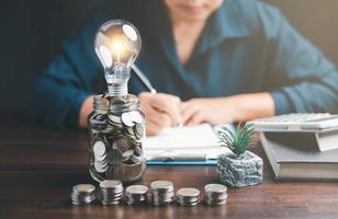 Businesswoman with lightbulb on money jar and using calculator to calculate and money stack. Save energy and money with accounting finance in office concept. Idea of energy saving planning in home. photo