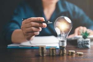 Businesswoman with lightbulb on coins and using calculator to calculate and money stack. Save energy and money with accounting finance in office concept. Idea of energy saving planning in home. photo