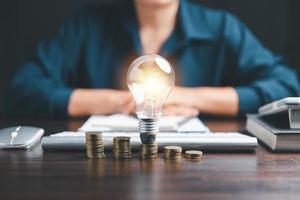 Businesswoman with lightbulb on coins and using calculator to calculate and money stack. Save energy and money with accounting finance in office concept. Idea of energy saving planning in home. photo