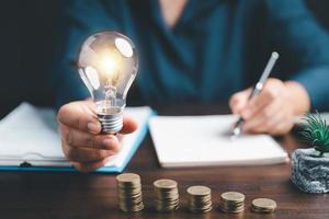 Businesswoman hand holding lightbulb with using calculator to calculate and money stack. Save energy and money with accounting finance in office concept. Idea of energy saving planning in home. photo