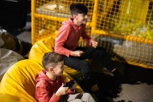 dos hermanos jugando vídeo juego consola, sentado en amarillo puff en niños jugar centro. foto