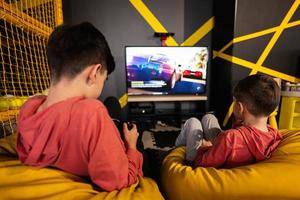 Two brothers playing race video game console, sitting on yellow pouf in kids play center. photo