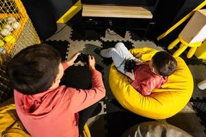 Two brothers playing video game console, sitting on yellow pouf in kids play center. photo