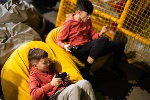 dos hermanos jugando vídeo juego consola, sentado en amarillo puff en niños jugar centro. foto