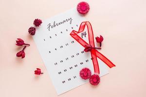 San Valentín día. un rojo en forma de corazon cinta Destacar el fecha febrero 14 en un calendario sábana y seco flores en un rosado antecedentes. parte superior ver foto