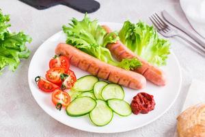 Listo a comer frito salchichas, Cortado Tomates y Pepino vegetales en un plato y un bollo en el mesa. abundante hecho en casa bocadillo foto