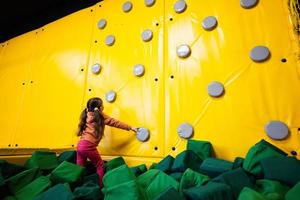 pequeño niña niño alpinismo pared a amarillo patio de recreo parque. niño en movimiento durante activo entretenimientos foto