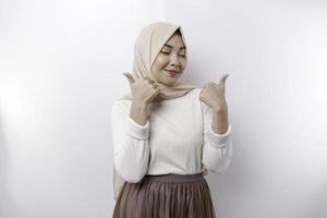 Excited Asian Muslim woman wearing a headscarf gives thumbs up hand gesture of approval, isolated by white background photo