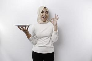 A smiling Asian Muslim woman is fasting and hungry and holding and pointing to a plate photo