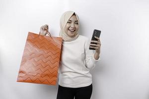 Portrait Asian Muslim woman standing excited holding an online shopping bag and her smartphone, studio shot isolated on white background photo