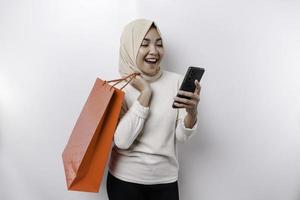 Portrait Asian Muslim woman standing excited holding an online shopping bag and her smartphone, studio shot isolated on white background photo