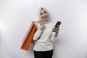 Portrait Asian Muslim woman standing excited holding an online shopping bag and her smartphone, studio shot isolated on white background photo